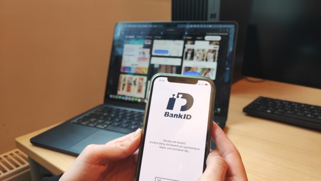 I fokus av bilden håller någon i en telefon med bank-ID uppe. I bakgrunden syns en laptop med en nätbutik.