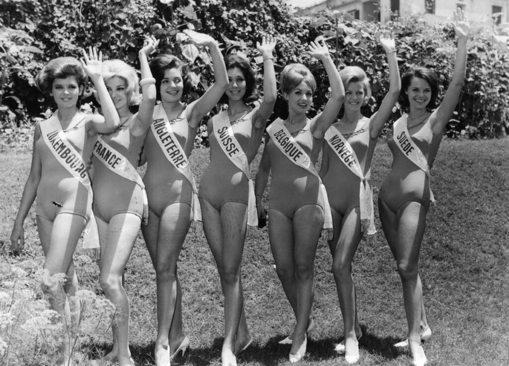 Sju modeller från en Miss Europe-tävling står bredvid varandra. Alla ler och vinkar. Det är 60-tal och bilden är svartvit.