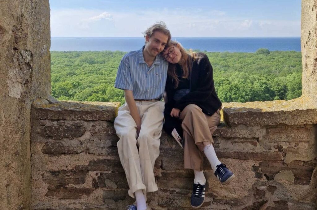 Bild på Max och Elvira som sitter på en gammal mur, på en kulle högt upp med havet långt bort i bakgrunden.