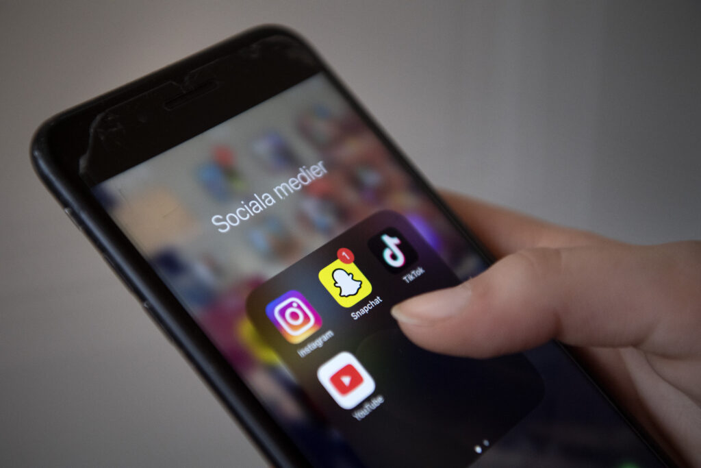Bilden föreställer en ung vuxen som håller i en mobiltelefon. Mobilen visar olika sociala medier. Instagram, Snapchat, TikTok och Youtube.